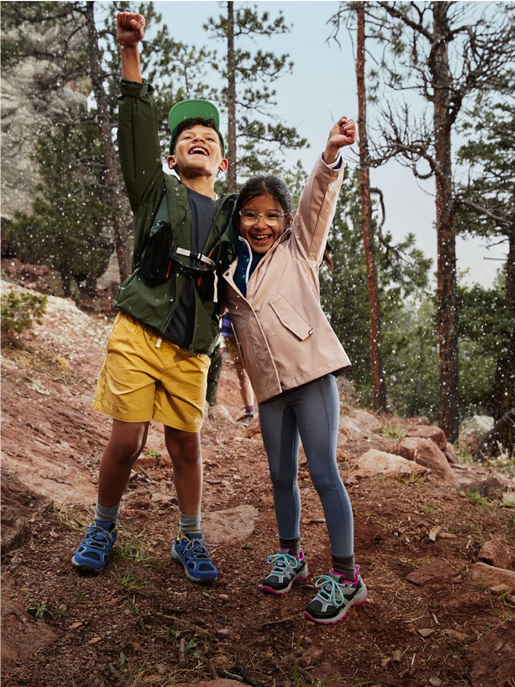 Two kids hiking in Merrell shoes smiling and raising their arms in the forest.